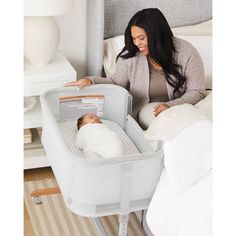 a woman laying in bed next to a baby in a bassinet and changing table