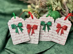 three christmas tags with bows on them sitting next to ornaments and greenery in the background