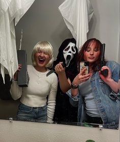 two women standing in front of a mirror holding up cell phones and wearing scary masks