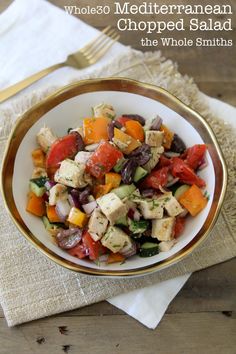 a white bowl filled with lots of food next to a fork and napkin on top of a wooden table