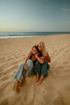 two women are sitting on the sand at the beach and one is hugging her friend