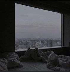 an unmade bed in front of a large window with cityscape behind it