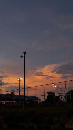 the sun is setting behind a fence and street lights in front of a building with a chain link fence