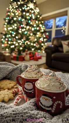 two mugs with whipped cream and cookies on a blanket in front of a christmas tree