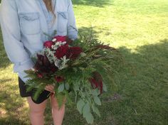 a woman standing in the grass holding a bouquet of red and white flowers with greenery