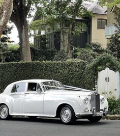 an old fashioned white car parked in front of a house with hedges on the side