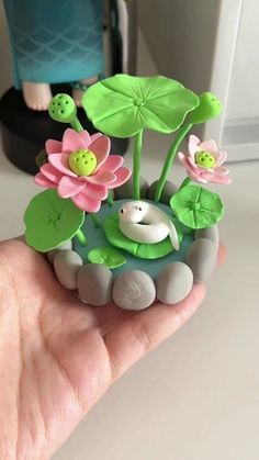 a hand is holding a small toy pond with flowers and rocks on it, while a doll stands in the background