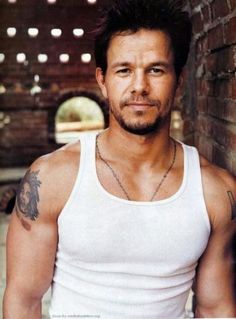 a man wearing a white tank top standing in front of a brick wall and looking at the camera