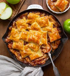 a skillet filled with apple pie next to two green apples on a wooden table