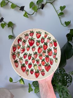 a hand holding a plate with strawberries on it and green plants in the background