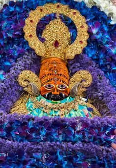 an elaborately decorated statue in the middle of purple and blue flowers, with a gold mask on it's head