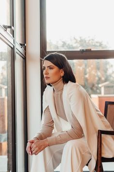 a woman sitting on a chair in front of a window wearing a white suit and heels