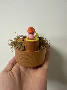 a hand holding a small wooden box with an orange and pink doll in it's center