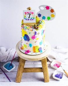 a colorful birthday cake sitting on top of a wooden stool next to paintbrushes