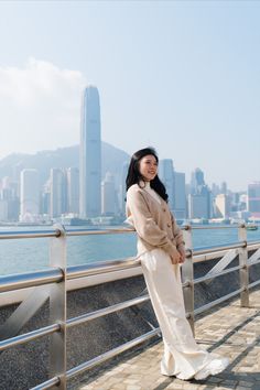 a woman standing on a bridge in front of the water and cityscape behind her