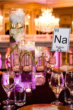 the table is set with wine glasses, candles and flowers in tall glass vases