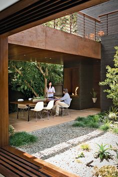 two people are sitting at a table in the backyard with plants and rocks on the ground