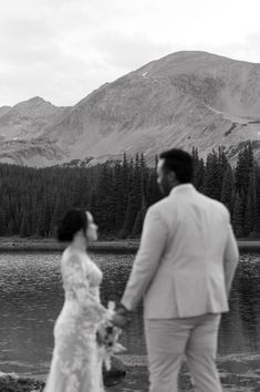 a bride and groom standing next to a lake with mountains in the background on their wedding day