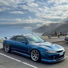 a blue sports car parked in a parking lot next to the ocean and people sitting on benches