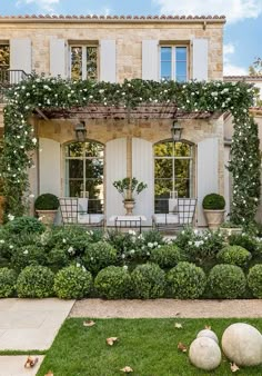 a house with white shutters and flowers in the front yard, surrounded by greenery