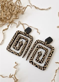 two square shaped wooden earrings with black and white designs on the sides, sitting next to dried grass