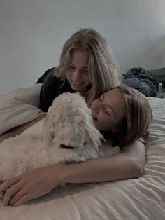 two women laying on a bed cuddling with a white dog and smiling at the camera