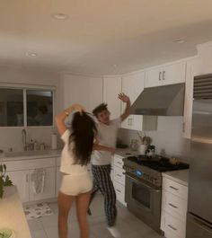 two people dancing in a kitchen with white cabinets and stainless steel stove top ovens