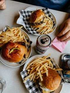 three plates with hamburgers and french fries on them next to two cans of soda