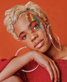 a woman with bright makeup and colorful face paint poses for a magazine cover shot in front of an orange background