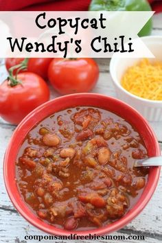 a red bowl filled with chili next to some tomatoes and other ingredients on a white wooden table