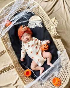 a baby is laying in a basket with pumpkins on it's head and wearing a knitted hat