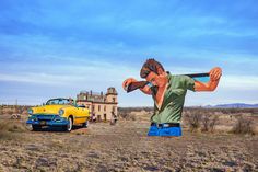 a man standing next to a yellow car on top of a dirt field in front of a building