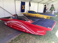 two people standing next to a red and yellow race car on display in a tent