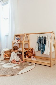 a toddler sitting on the floor in front of a shelf with clothes and toys