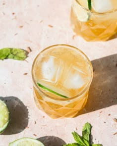 two glasses filled with ice and limes on top of a pink countertop next to mint leaves