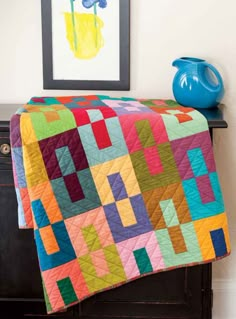 a bed with a colorful quilt on top of it next to a blue vase and painting