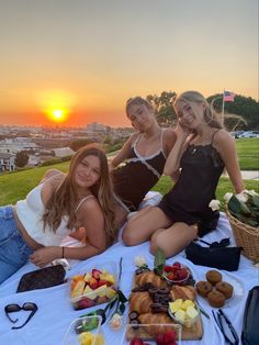 three beautiful women sitting on top of a white table covered in fruit and pastries