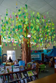 a library filled with lots of books under a tree covered in colorful paper leaves and hanging from the ceiling