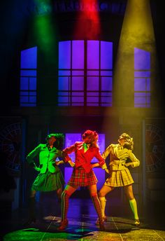 three women in plaid skirts and high heels are performing on stage with bright colored lights behind them