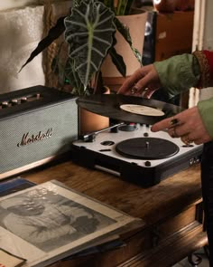 a person standing next to a record player on top of a wooden table in front of a potted plant