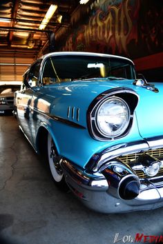 an old blue car parked in a garage with graffiti on the wall behind it's headlight