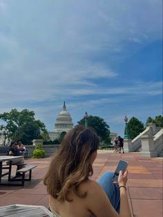 a woman sitting on a bench looking at her cell phone in front of the capitol building