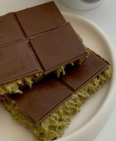 two pieces of chocolate and green food on a white plate with a cup of coffee in the background