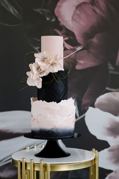 a black and white cake with pink flowers on top sitting on a gold stand in front of a floral wallpaper