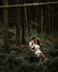 a man and woman sitting on a swing in the middle of a forest with trees