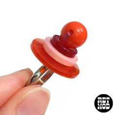 a hand holding a red and pink button on top of a metal ring with an orange ball attached to it