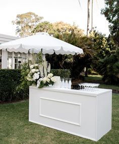 a white bar with flowers and wine glasses under an umbrella on the grass in front of a house