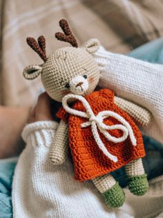 a hand holding a stuffed animal wearing an orange sweater and green pants with deer antlers on it's head