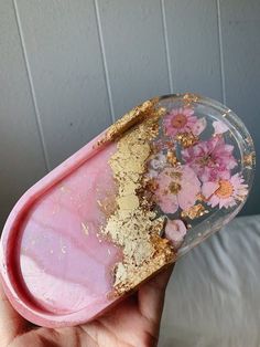 a hand holding a pink and gold tray with flowers on the inside, in front of a white wall