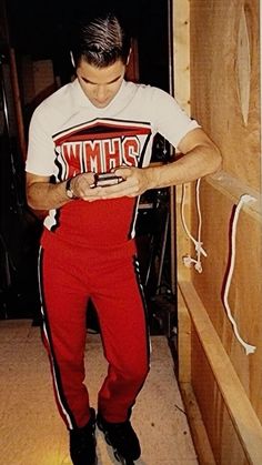 a man in red pants looking at his cell phone while standing next to a wooden wall